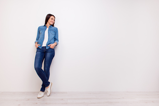 Full-length portrait of beautiful smiling woman holding hands in pockets and leaning on white wall