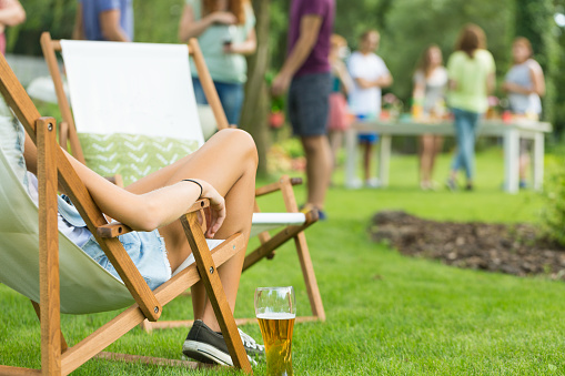 Close-up of beautiful young woman's legs, girl enjoying summer vacation, relaxing while sitting on deck chair with beer, friends at garden party in the background
