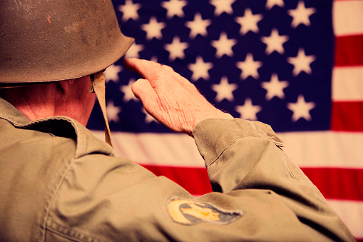 Senior adult man in military combat uniform saluting the American flag.  Rear view.