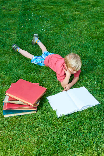 Child boy reading book, lying down on green grass. Blank pages for your content