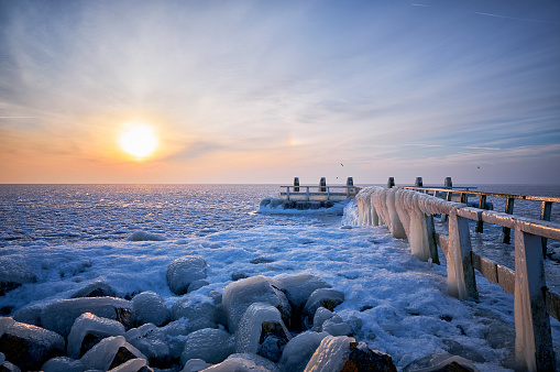 In the early morning at a frozen lake during sunrise. The jetty and its poles are covered with ice due to the water which had been blown on the jetty and poles by wind force before the lake was frozen.