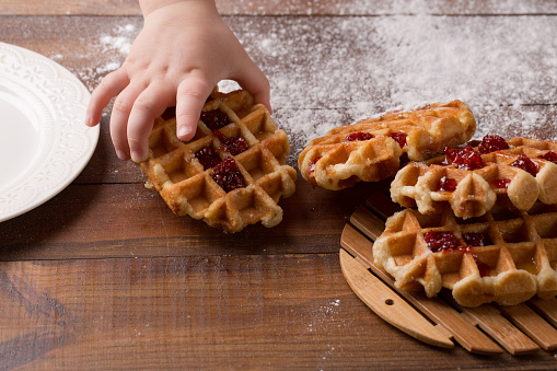 child's hand takes Tasty homemade waffle and strawberry jam. wooden background with scattered flour