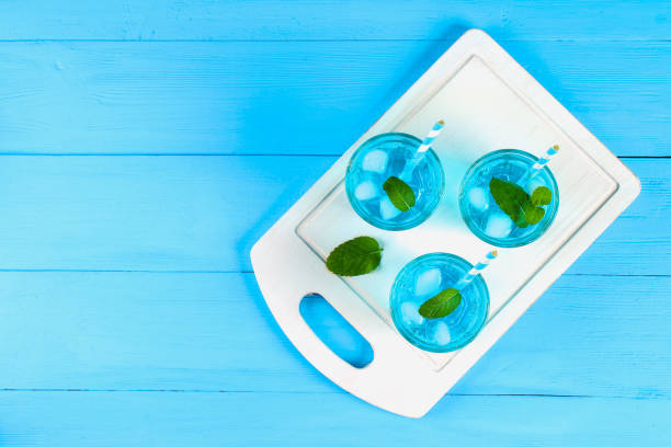 istock Blue cocktail with ice and mint in glasses on a white wooden board on a blue table. Top view. 939772490