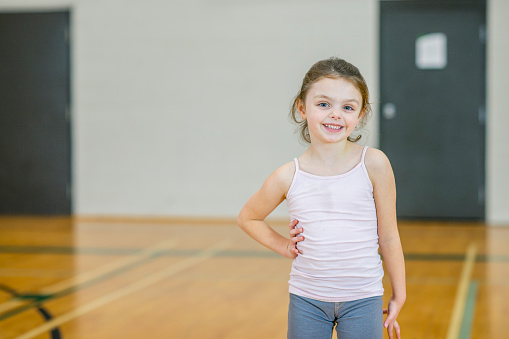 An adorable caucasian 6-7 year old girl has her hand on her hip and smiles in gym class.