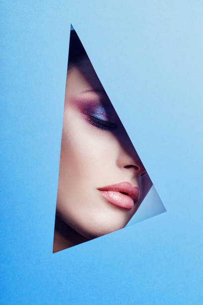 istock Woman with a beautiful bright makeup and pink lipstick looks through a triangular hole in the blue paper. Advertising cosmetics, professional art makeup, lip gloss, bright color, blue background 939827202