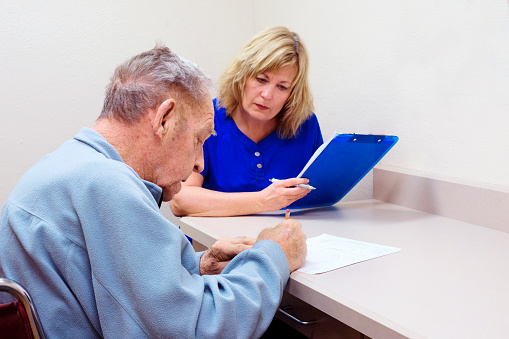 Blond Caucasian speech language pathologist evaluates the cognitive-communication skills of a senior aged man in a clinic setting who is completing the clock-drawing test.