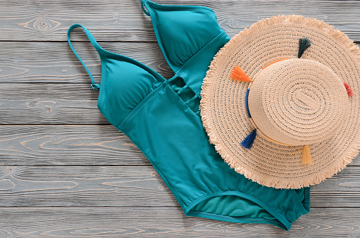 Womens clothing, accessories (straw hat, blue green swimsuit) on grey wooden background with copy space. Trendy fashion outfit. Shopping, travel, summer, beach concept, abstract.  Flat lay