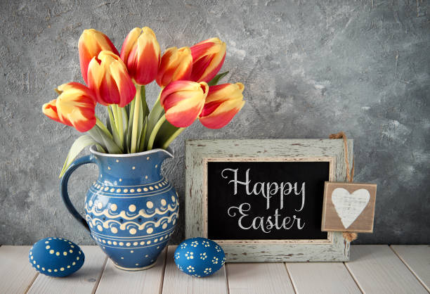 istock Red-yellow tulips in blue ceramic pitcher with Easter eggs and a blackboard on gray background, text 939892162