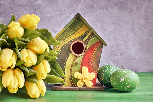 Easter decorations on gray concrete background. Yellow tulips, birdhouse and painted Easter eggs with decorative felt flower