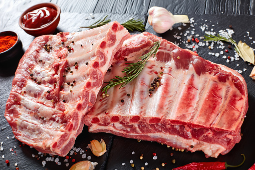 fresh rack of raw pork spare ribs seasoned with spices on black slate tray with paprika, garlic cloves, bay leaves at background, vertical view from above, close-up