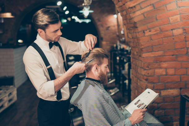 istock Red bearded guy is reading a magazine in a barber shop while getting bren new haircut from a classy dressed stylist. Both are buzy 939989620
