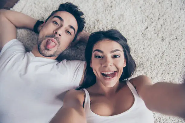 Photo of Cheerful playful hispanic sister and brother are taking selfie and making funny grimaces. They lie on the floor on beige carpet in white casual outfits indoors at home