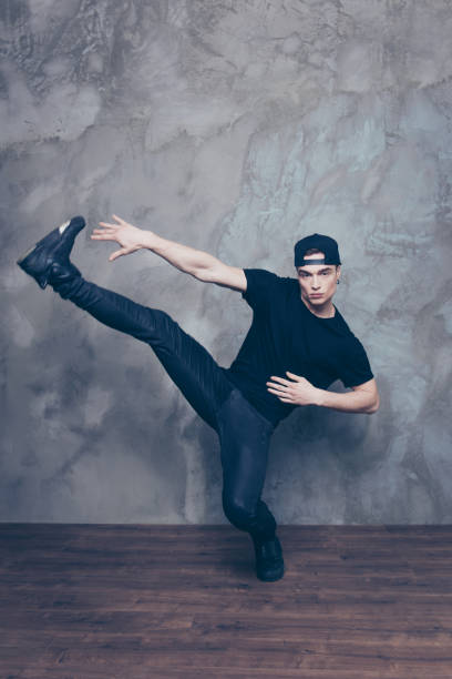 istock Handsome young break dancer is making his favourite move in studio on the grey background. He is wearing black stylish outfit and cap 940006334