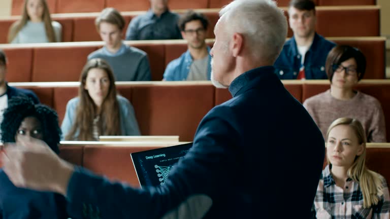 Back View of the Professor Giving Lecture on Computer Science to a Classroom Full of Multi Ethnic Students. Teacher Holds Laptop with Deep Learning, Artificial Intelligence Infographics on the Screen.