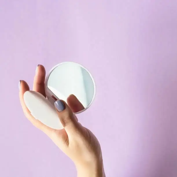 A compact, compact mirror in women's hands. On a lilac bright background. Makeup female accessories