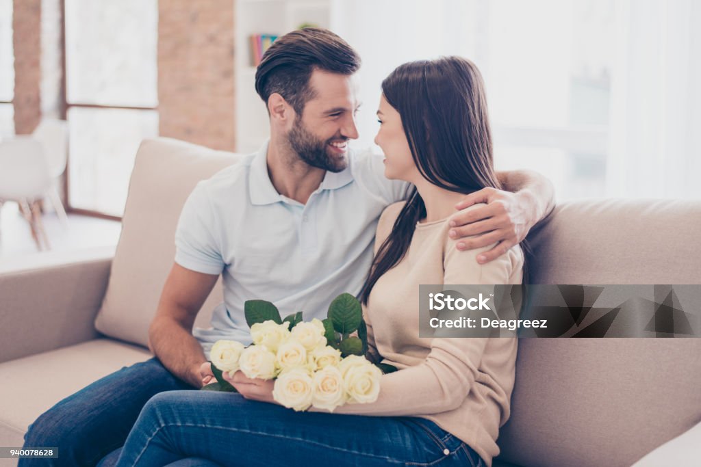 Happy together. Man is embracing his girlfriend with roses in hands. They are on sofa at home Couple - Relationship Stock Photo