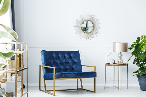 Navy blue armchair next to gold table with lamp against white wall with mirror in living room interior