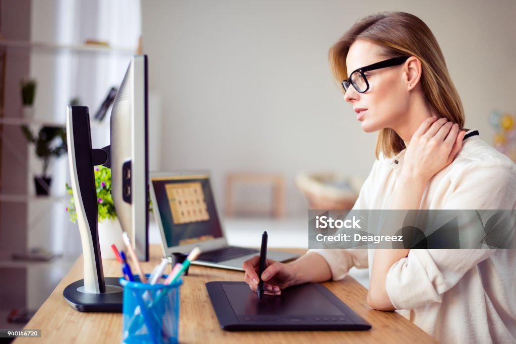 Concentrated young woman in glasses retouching photo for a long time and having  pain in her neck Computer Monitor Stock Photo