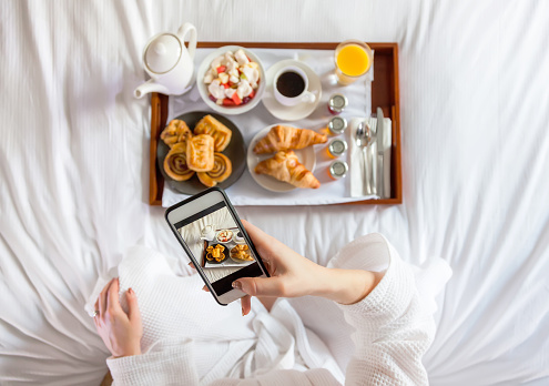 Young woman who wears bathrobe taking photo of breakfast by mobile phone in bed at the hotel room.