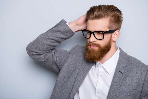 Confident pensive handsome bearded young man is looking away wearing suit and glasses. fixing his perfect hairstyle
