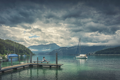 A woman sits on wooden pier by a lake (Lucerne Lake, Switzerland), as rain clouds gather above.