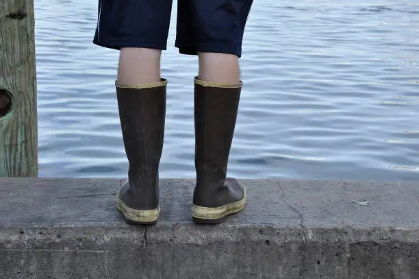 A picture of a boy from the waist down sporting rubber boots & shorts is about to go fishing