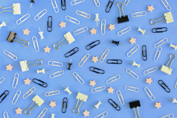 istock Stationery chaotically scattered on a blue background. White and black paper clips, clerical buttons and small pink stars in the riots. Picture from the top. 940284364
