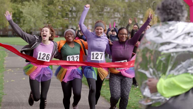 Playful women friend runners in tutus crossing finish line at charity fun run