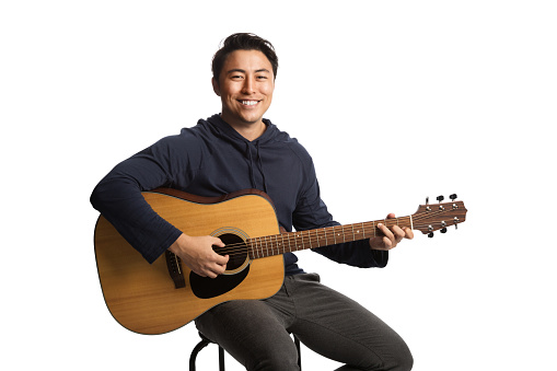 Smiling young man wearing a blue hoodie strumming on a acoustic guitar. Sitting down in front of a white background.