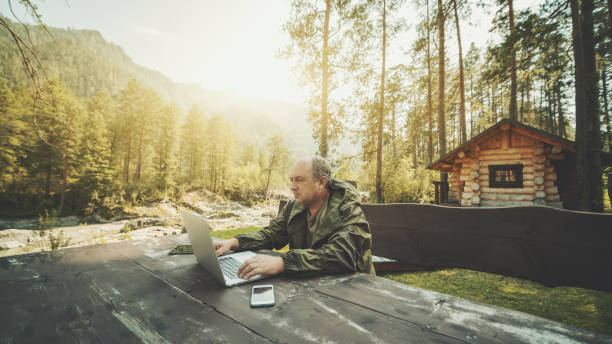 Man entrepreneur near his summer house with the laptop Adult partly bald hunter is sitting outdoors near his shack at the wooden table and working on his laptop; aged businessman in overalls is working remotely on the netbook during his vacations altai nature reserve photos stock pictures, royalty-free photos & images