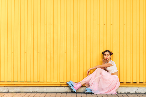 young girl in pink skirt and roller skates sits on the sidewalk against the background of yellow wall and makes an air kiss. Concept life style. Copy space.