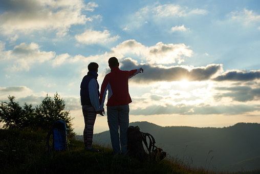 Silhouette of two tourists are standing on top of the mountain holding hands near backpacks against the backdrop of mountains and cloudy sky at sunset. Guy showing his hand in the distance. Rear view