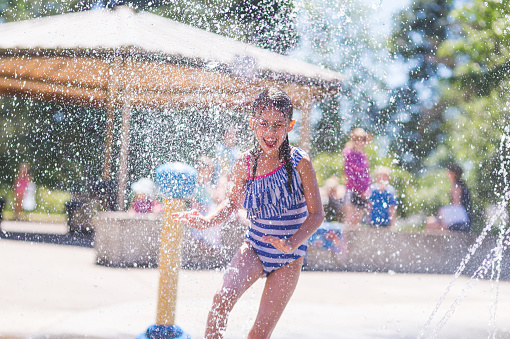 A young ethnic girl splashes and plays in a playground splash pad. The water is cascading all around her and she is smiling as she runs through. It is a sunny, perfect day for getting wet and playing hard!