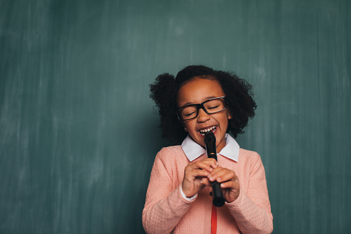 A young nerd girl dressed in sweater, eyeglasses plays her recorder. She has a fun smile on her face and is ready to become a master of music. Music is cool.