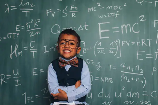A young nerd boy dressed in sweater, eyeglasses sits in front of a chalkboard full of math equations in a school classroom. He has an inquisitive smile on hisr face and is ready to excel in academics.