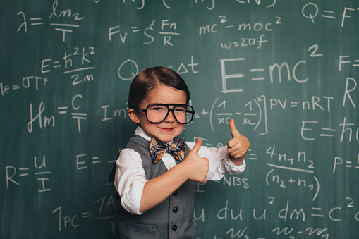 A young nerd boy dressed as a professor in sweater, bow tie and eyeglasses sits in front of a chalkboard full of math equations in a school classroom. He has a happy smile on his face giving thumbs up and is ready to teach academics.