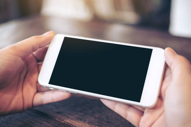 Mockup image of hands holding white mobile phone with blank black screen for watching and playing games on wooden table in cafe
