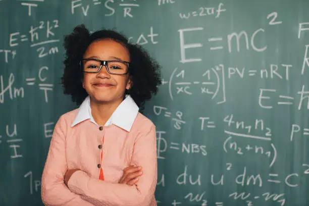A young nerd girl dressed in sweater, eyeglasses sits in front of a chalkboard full of math equations in a school classroom. She has an inquisitive smile on her face and is ready to excel in academics.