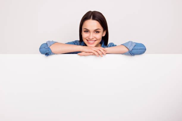 pretty smiling woman in jeans shirt leaning on the top of the white banner,  place for copy-space on board - advertisement advertise businesswoman peeking imagens e fotografias de stock