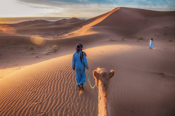 Showing the traditional blue clothing and mode of transportation of nomadic Tuareg tribesmen in the Sahara Desert, as they walk through the sand dunes with camels. stock photo