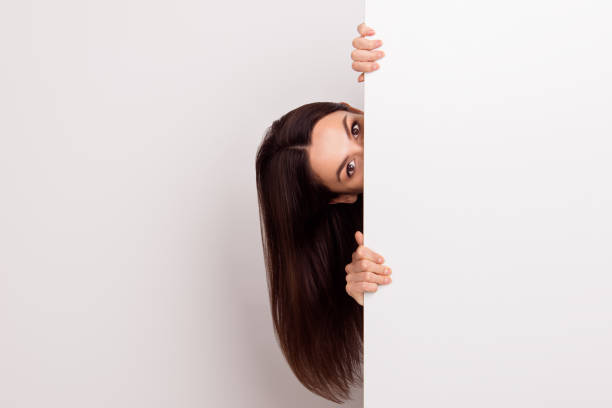 cute young woman with long hair peering around from behind white board, place for copy-space - advertisement advertise businesswoman peeking imagens e fotografias de stock