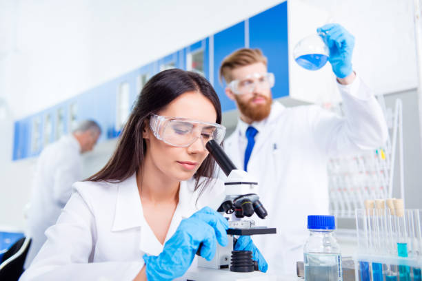 istock Team work. Young lab worker in safety glasses is analysing the sample in the microscope. She is in a labcoat, in the middle of experiment 940378612