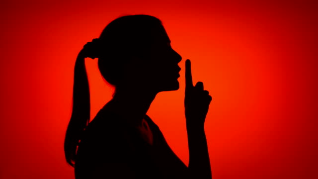 Silhouette of young woman making silence gesture on red background. Concept of mystery and secrecy
