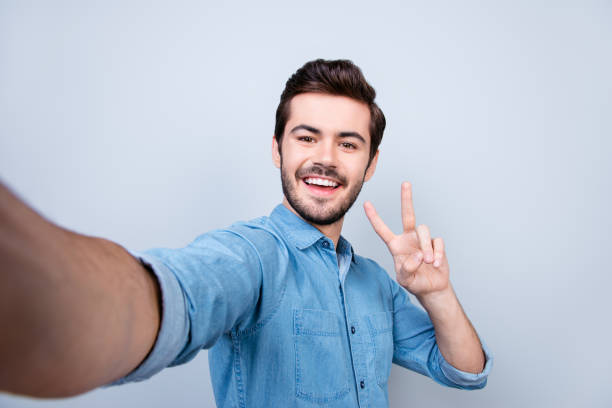 istock Handsome smiling young man is making selfie on smartphone and gesturing peace on light blue background 940462214