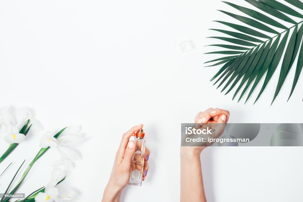 Top view of female hands with pink manicure Top view of female hands with pink manicure spraying perfume on wrist among the palm branch and iris flowers on white background. Stylish flat lay frame with copy space for text. Perfume Stock Photo