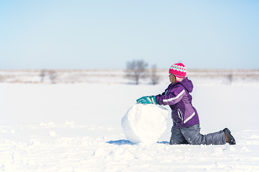 Side view of a young girl rolling a large snowball for a snowman. She is on a frozen lake covered in snow on a late winter/early springtime day in Minnesota.