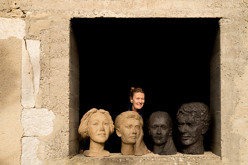 Natural portrait of a sculptor who lives and works in la Drome, Provence, France. She is standing behind clay busts that she has made. The head on the left is a self-portrait.
