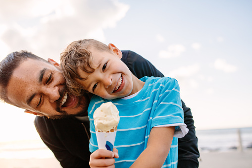 Boy eating an ice cream standing near seafront with his father. Little boy holding an ice cream cone while his father playfully tries to eat it from behind.