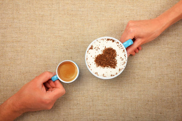 Close up man and woman hands hold small espresso and big full cup of latte cappuccino coffee with heart shaped chocolate on milk froth over canvas tablecloth, elevated top view, directly above