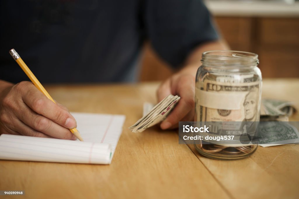 Counting Cash at the Kitchen Table A male writes notes concerning money at the kitchen table.  On the table is a pencil, pad of paper, paper currency and a jar fill with cash and coins.  The jar as blank masking tape to hold text. Jar Stock Photo